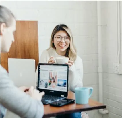 A casual office setting where two people are sitting at a table. The woman on the right is smiling and holding a coffee cup. The man on the left, facing away from the camera, is working on a laptop. The laptop screen displays a webpage or application. The setting appears relaxed, with natural light and simple decor